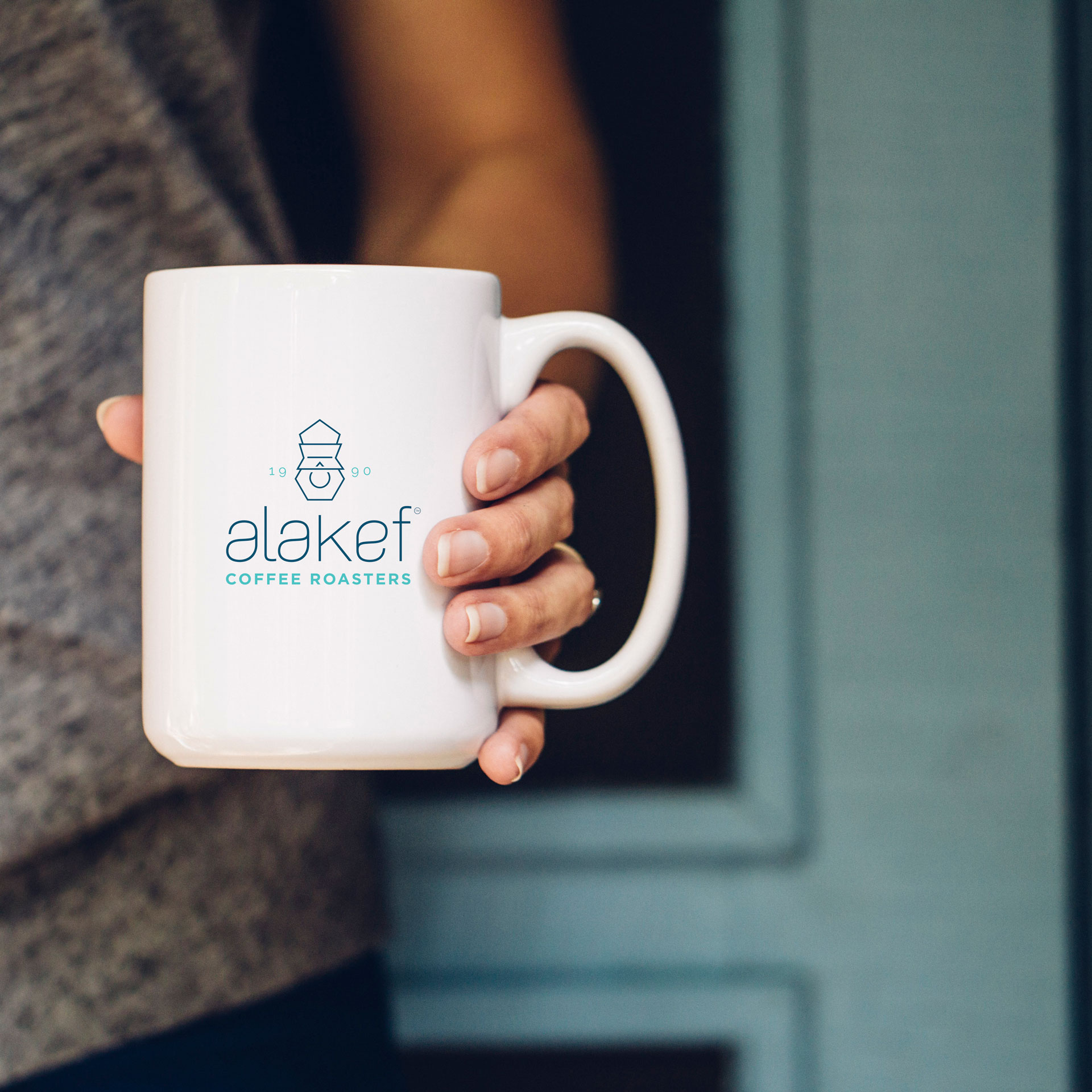 A person holds a white coffee mug with the logo and text "alakaf COFFEE ROASTERS" printed in blue. The background is slightly out of focus, showcasing a wooden door. The individual, wearing a gray shirt, enjoys their alakef coffee moment.