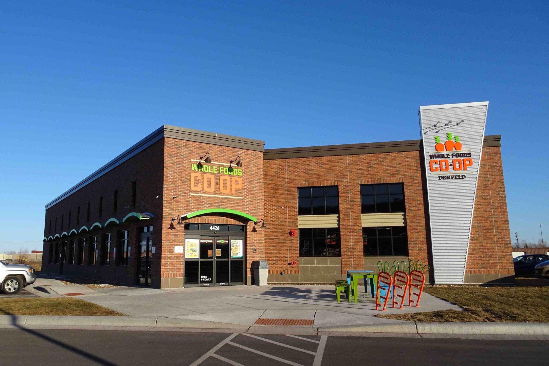 A brick building with signs reading "Whole Foods Co-op" above the entrance and on a tall panel stands proudly in Duluth, MN. The building features large windows and a bike rack out front. The entrance has double glass doors and some overhanging awnings, set against a clear sky backdrop.