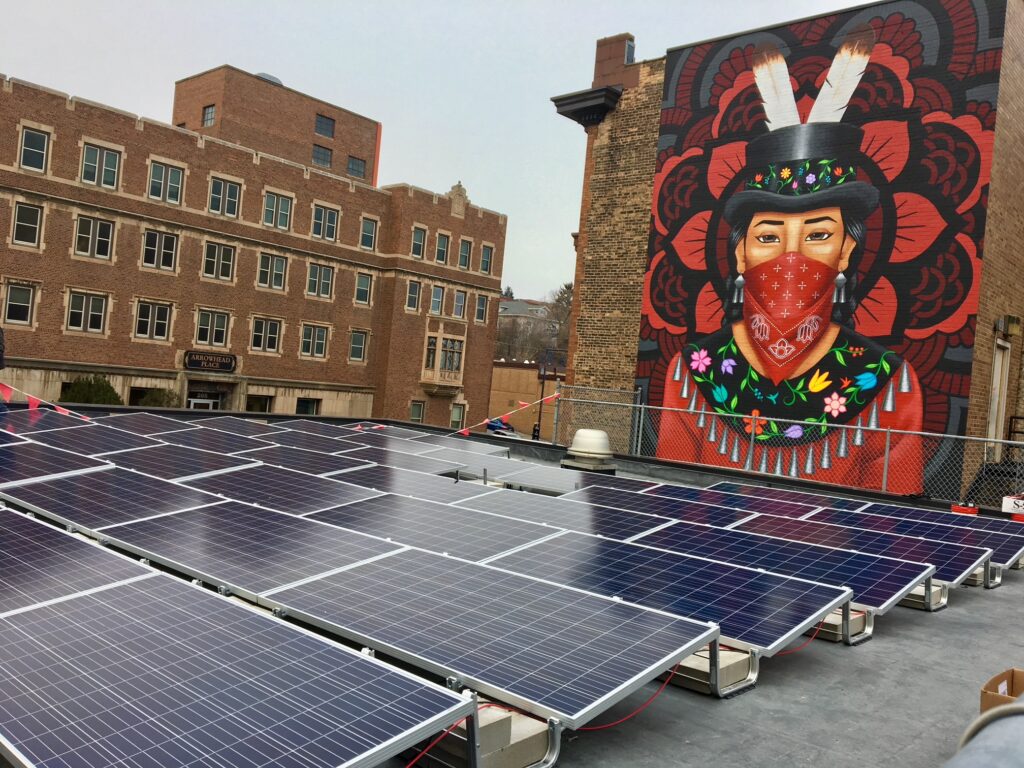 A rooftop with several solar panels in the foreground. In the background, there is a mural painted on a building featuring a person wearing a colorful headdress and a red bandana mask. Other brick buildings, perhaps hiding cozy nooks for coffee in Duluth, are visible on an overcast day.