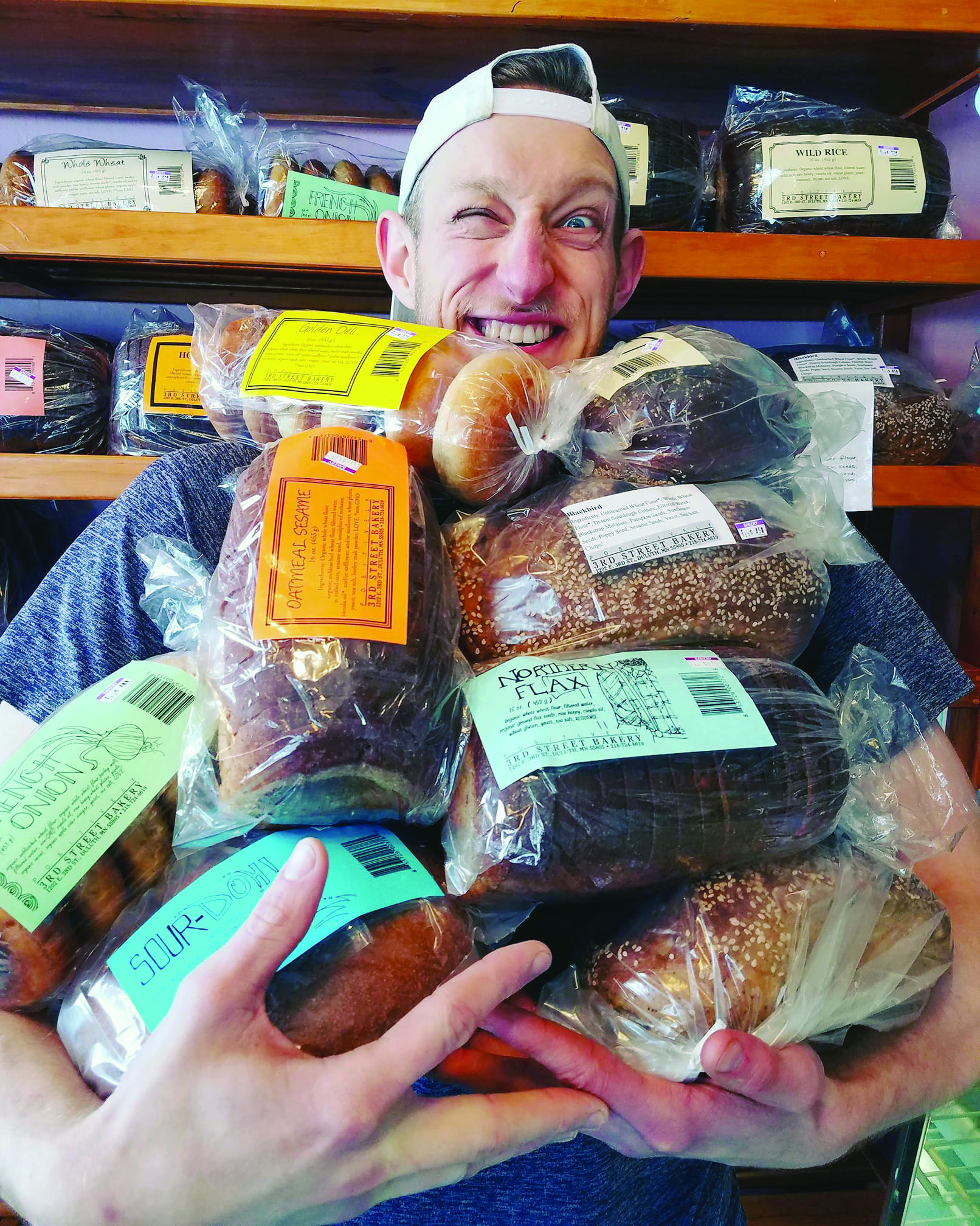 A person wearing a white cap backward is smiling while holding a large assortment of packaged bread. The background shows shelves filled with more bread, making it a perfect morning to enjoy some coffee in Duluth.