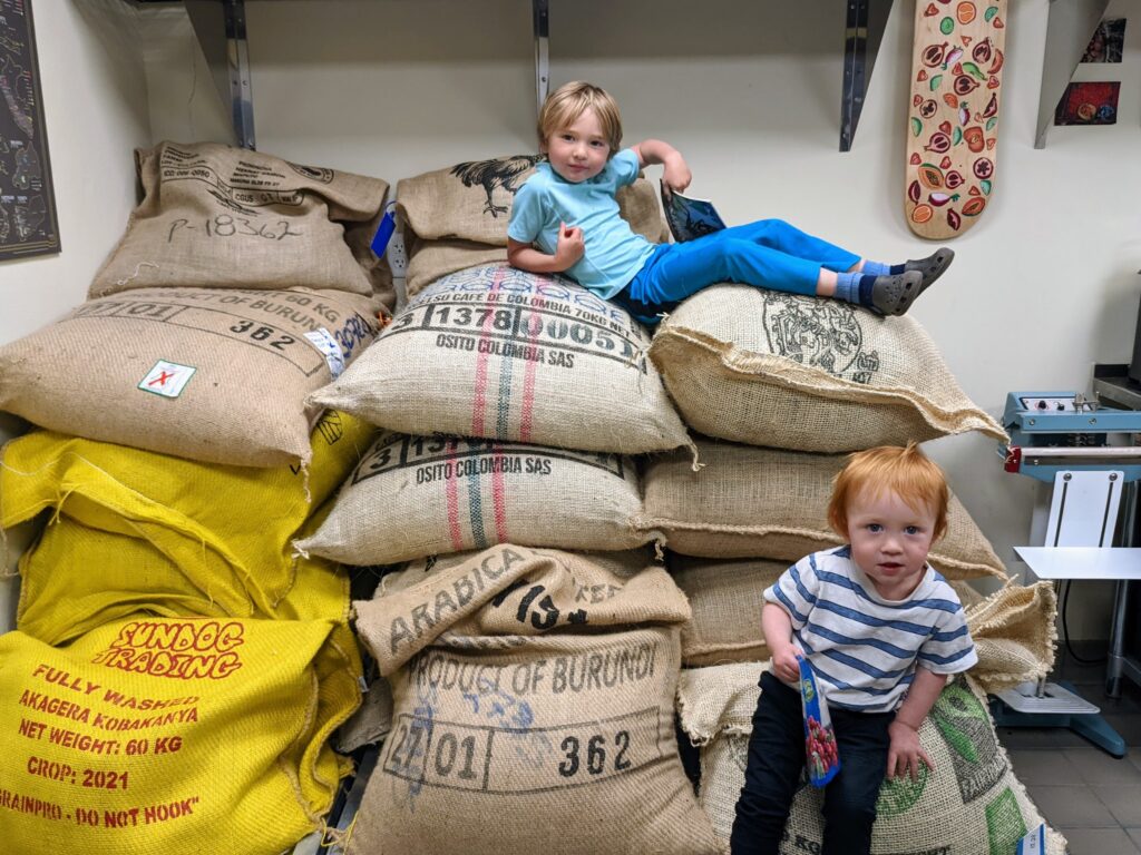 Two young children are sitting on a stack of large burlap coffee sacks in a room. The older child, with blond hair, is seated higher on the stack, while the younger child, with reddish hair, is lower. Both appear relaxed and curious. Shelves and a skateboard decorate the background wall, reminiscent of a cozy Coffee in Duluth vibe.