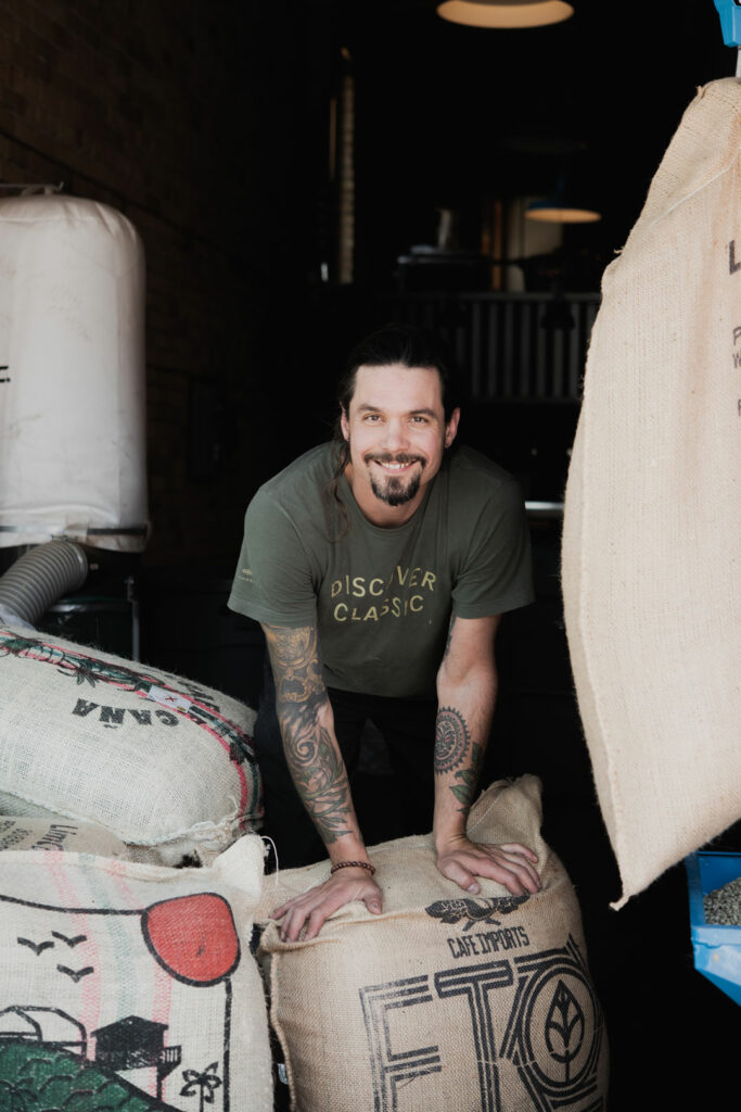 Man in front of a coffee roaster with bags of coffee around.