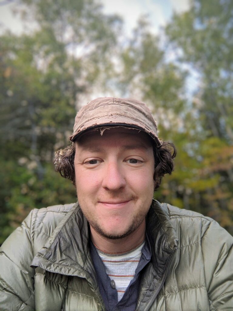 A person with curly hair, wearing a brown cap and green puffer jacket, smiling at the camera. The background features trees with green and yellow foliage, indicating a natural outdoor setting. The sky is partially visible and overcast, reminiscent of a cozy day spent enjoying coffee in Duluth.