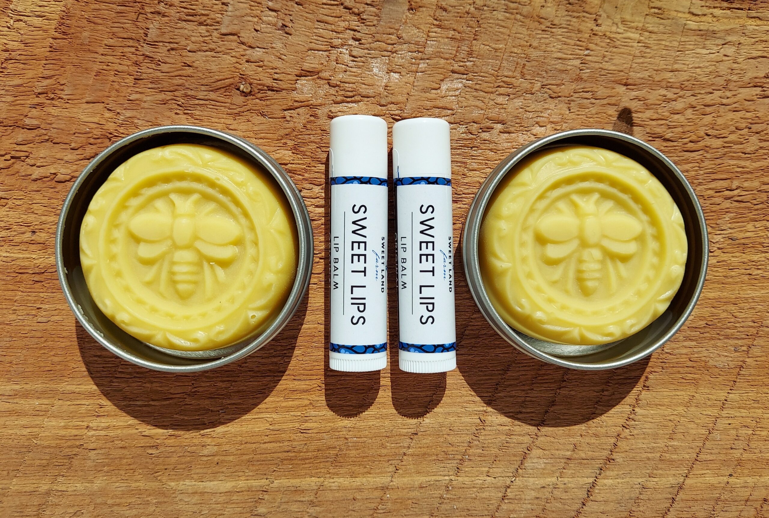 The image displays two round tins of solid lotion bars with bee imprints, placed on a wooden surface. Between the tins are two tubes of lip balm labeled "SWEET LIPS." The items are arranged symmetrically in bright sunlight, reminiscent of enjoying a perfect morning coffee in Duluth.