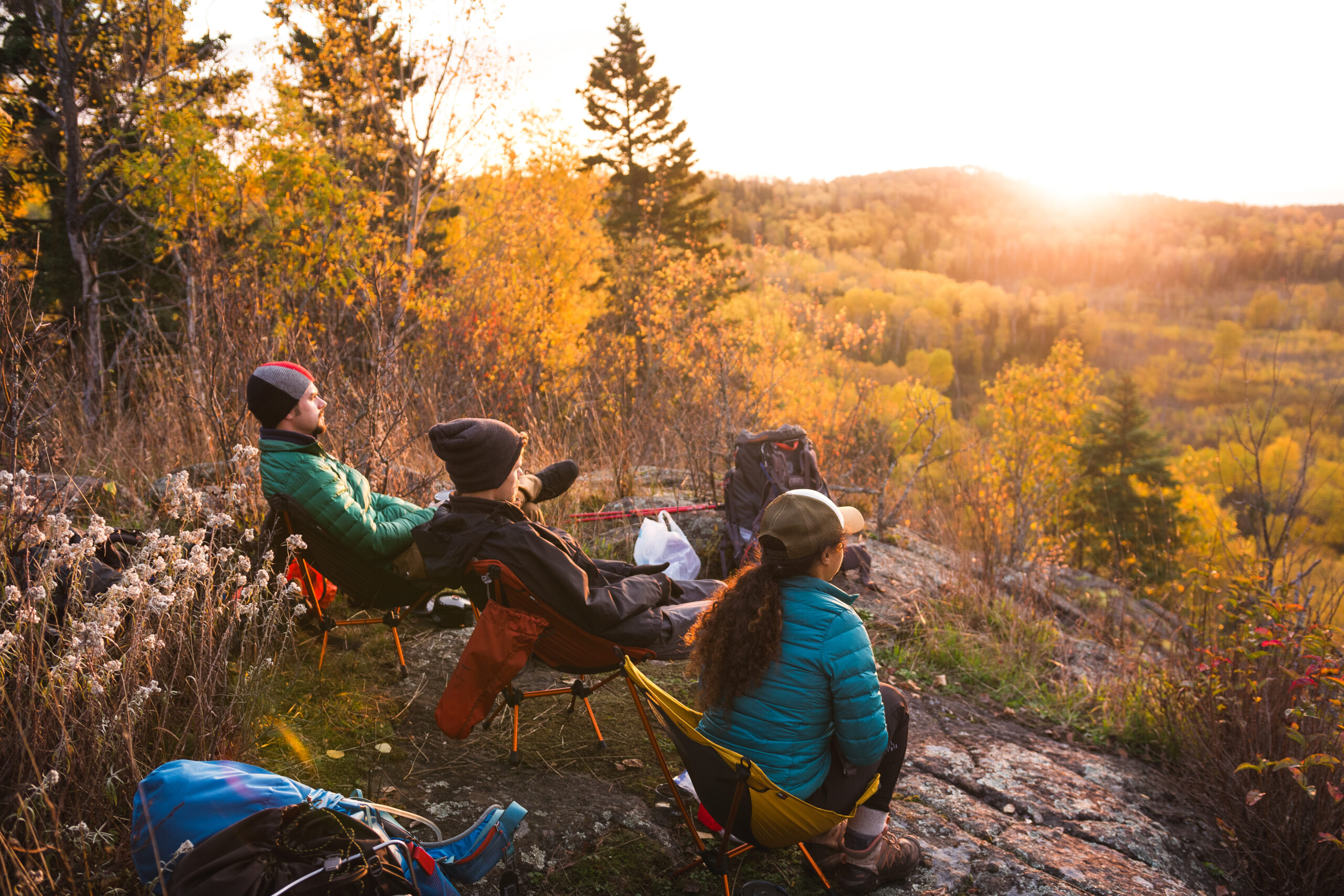 Three people sit on camping chairs atop a rocky cliff, overlooking a forest valley in autumn. They face a golden sunset, surrounded by trees with vibrant fall colors. Backpacks and gear are scattered around as they enjoy the serene natural view, like savoring coffee in Duluth on a crisp morning.