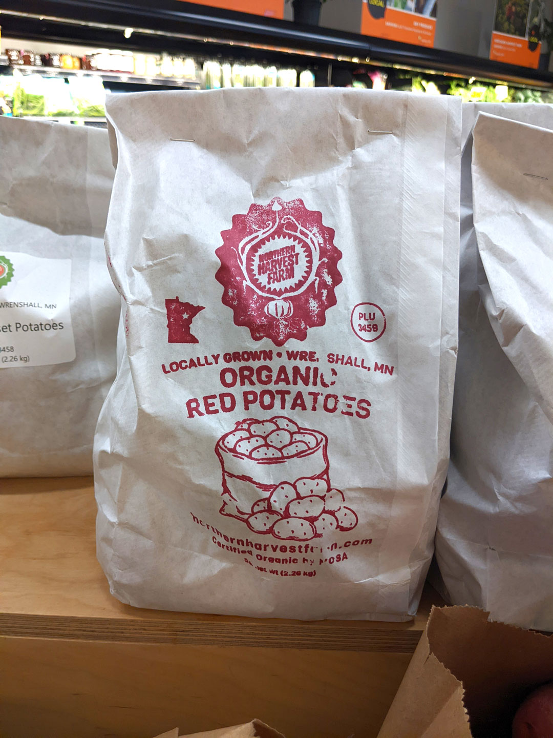 A paper bag labeled "Organic Red Potatoes" sits on a wooden surface. The bag features a red printed design with text stating "Locally Grown • WRE, Shall, MN" and "PLU 9489". The bag includes a logo with "Fresh Farm Harvest" and a website link.