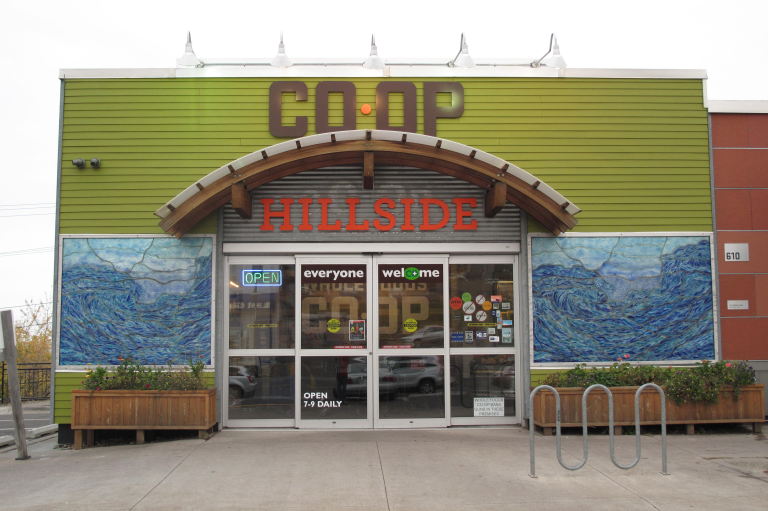 The entrance of a co-op store named "Hillside." The building's exterior is green with artistic murals on either side of the entrance. Signs on the glass doors display "Open," "everyone welcome," and operating hours. A bike rack is visible on the right.