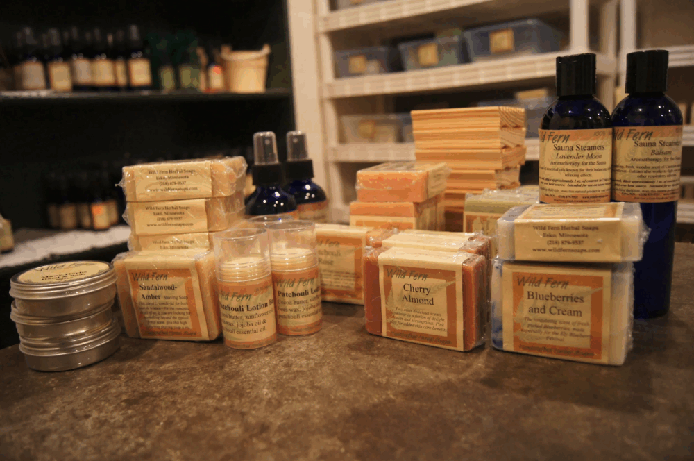 A variety of artisanal soaps, lotions, and skincare products are displayed on a wooden surface. The products include bars of soap labeled with scents like "Blueberries and Cream," "Cherry Almond," and "Amber." In the background, bottles and tins evoke the charm of a cozy coffee shop in Duluth.