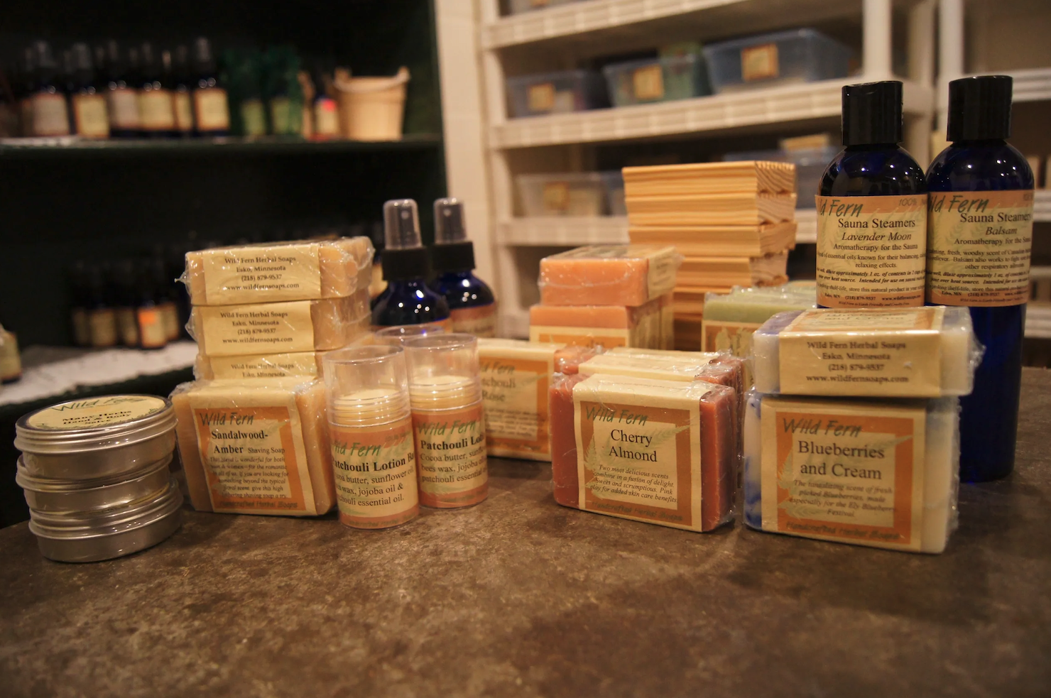 A variety of artisanal soaps, lotions, and skincare products are displayed on a wooden surface. The products include bars of soap labeled with scents like "Blueberries and Cream," "Cherry Almond," and "Amber." In the background, bottles and tins evoke the charm of a cozy coffee shop in Duluth.
