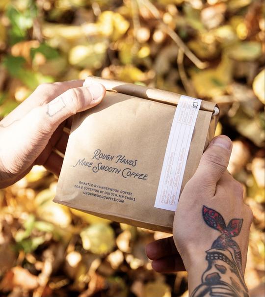 A person with a tattoo on their wrist holds a brown paper-wrapped coffee package labeled "Rough Hands Make Smooth Coffee." Sunlight filters through the autumn leaves in Duluth. The package, sourced locally, includes roasting details and contact information.