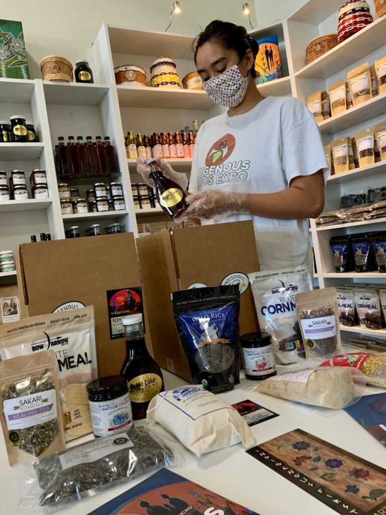 A person wearing a mask and gloves is packing various food items into a cardboard box in a store. Shelves filled with packaged goods and bottles stand behind them. Sporting a shirt that reads 