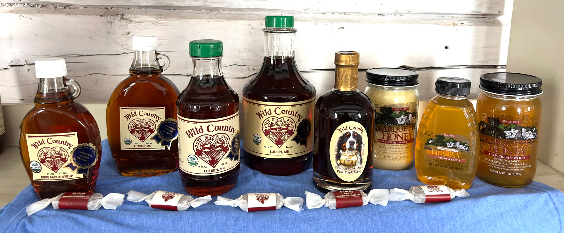An assortment of Wild Country maple syrup bottles and jars of honey are displayed on a blue cloth. The setup includes various syrup bottle shapes and sizes, honey jars, and wrapped candies, all arranged in front of a rustic white wooden background.