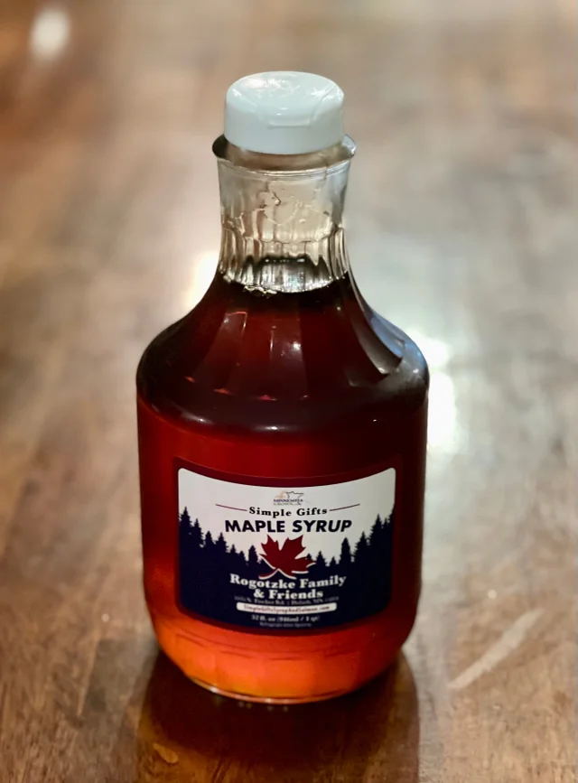 A glass bottle of "Simple Gifts Maple Syrup" sits on a wooden surface, reminiscent of enjoying coffee in Duluth. The syrup is dark amber in color. The label features a maple leaf graphic and reads "Rogatzke Family & Friends," with additional text. A white cap seals the bottle.