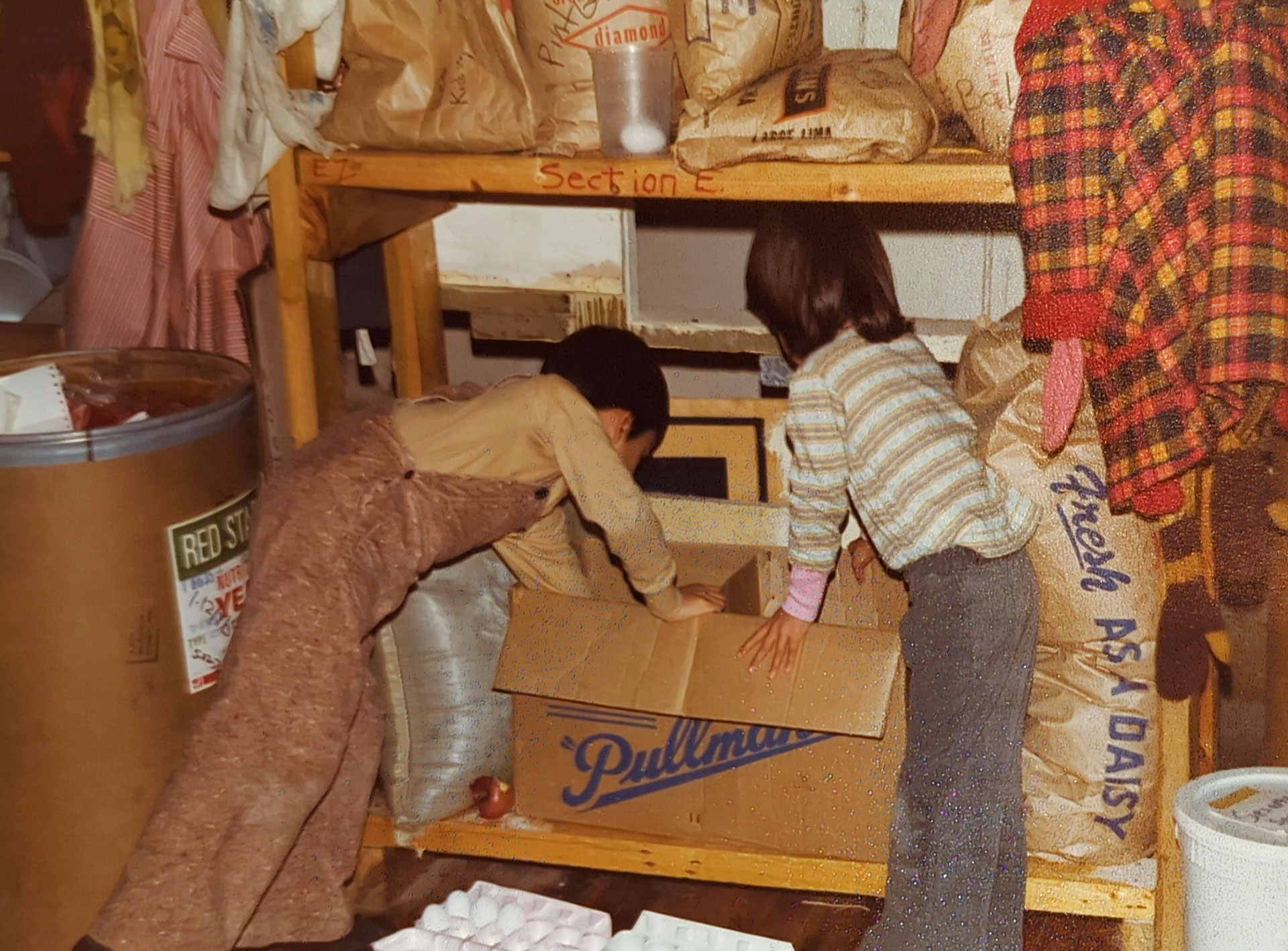 Two children are seen placing items into a cardboard box labeled "Pullman" on a wooden shelf in what appears to be a storage area reminiscent of the Whole Foods Coop Duluth MN. Surrounding them are large sacks of food and supplies. One child is wearing brown overalls and the other a striped sweater.
