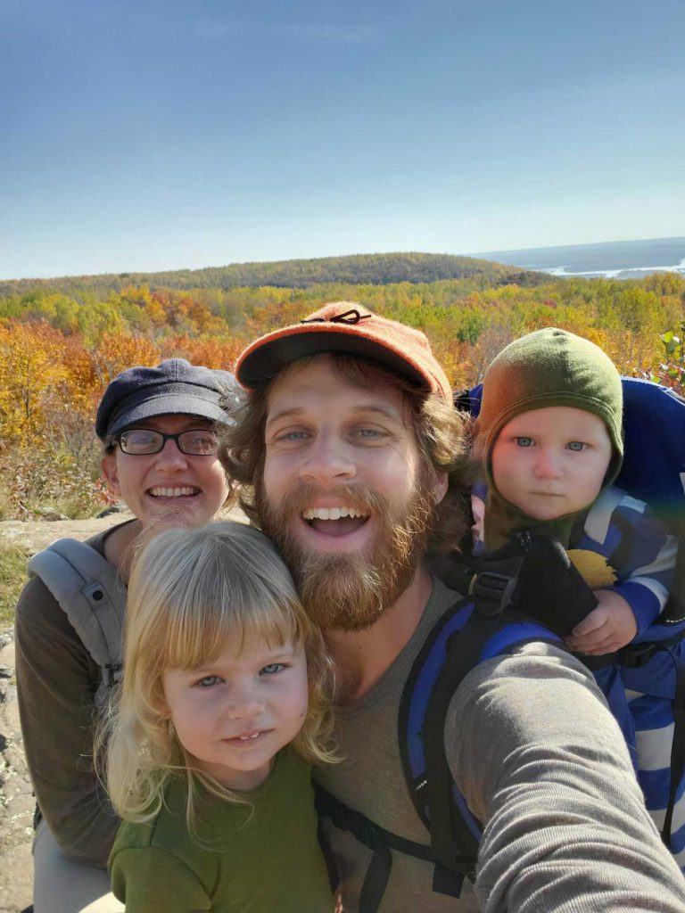 A family of four takes a selfie during a hike. The parents, wearing hats, smile at the camera while carrying two small children, one on the father's back in a carrier. The background features a scenic view of colorful fall foliage and a distant lake.