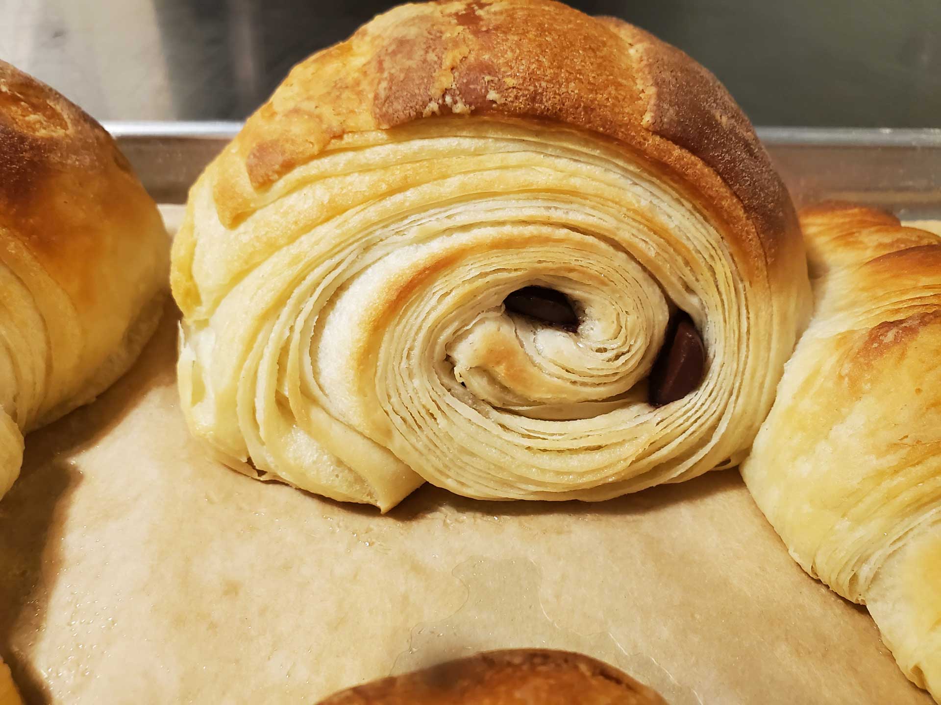 A close-up of a freshly baked, golden-brown croissant with visible layers of flaky pastry, resting on a parchment-lined baking sheet. The croissant has a swirl pattern in the center, revealing its intricate, buttery layers and a hint of chocolate filling.