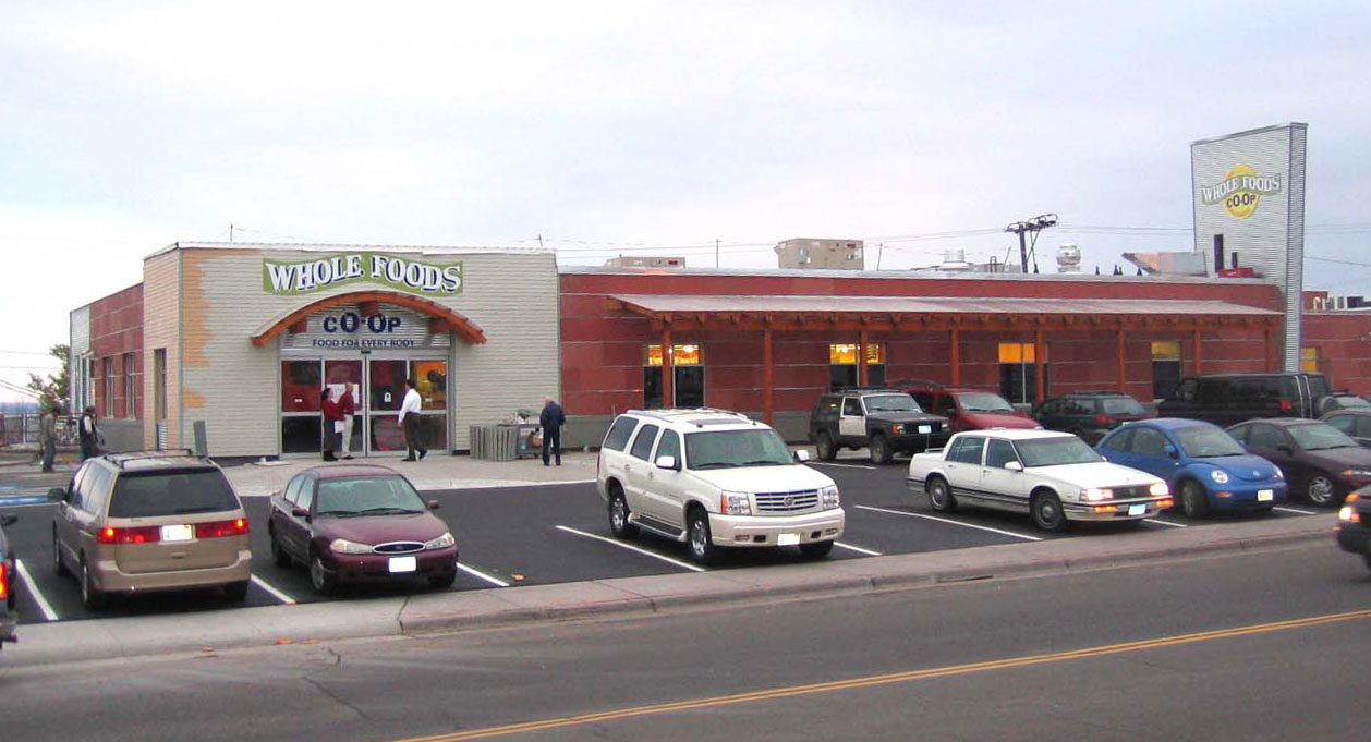 An exterior view of a Whole Foods Coop Duluth MN grocery store with cars parked in the lot. The store has a light brown facade with large windows and a sign above the entrance. The scene is peaceful with a clear sky and a few people entering and exiting the store.
