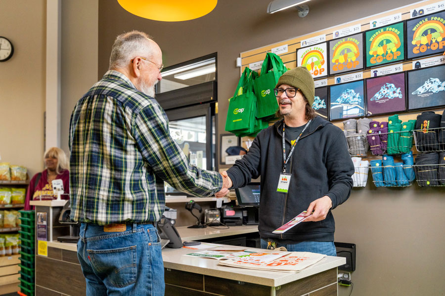 A man in a plaid shirt and a man in a beanie and glasses are shaking hands across a counter at the Whole Foods Coop in Duluth, MN. Shelves behind them display colorful products and reusable bags, while another person stands in the background near a rack of snacks.