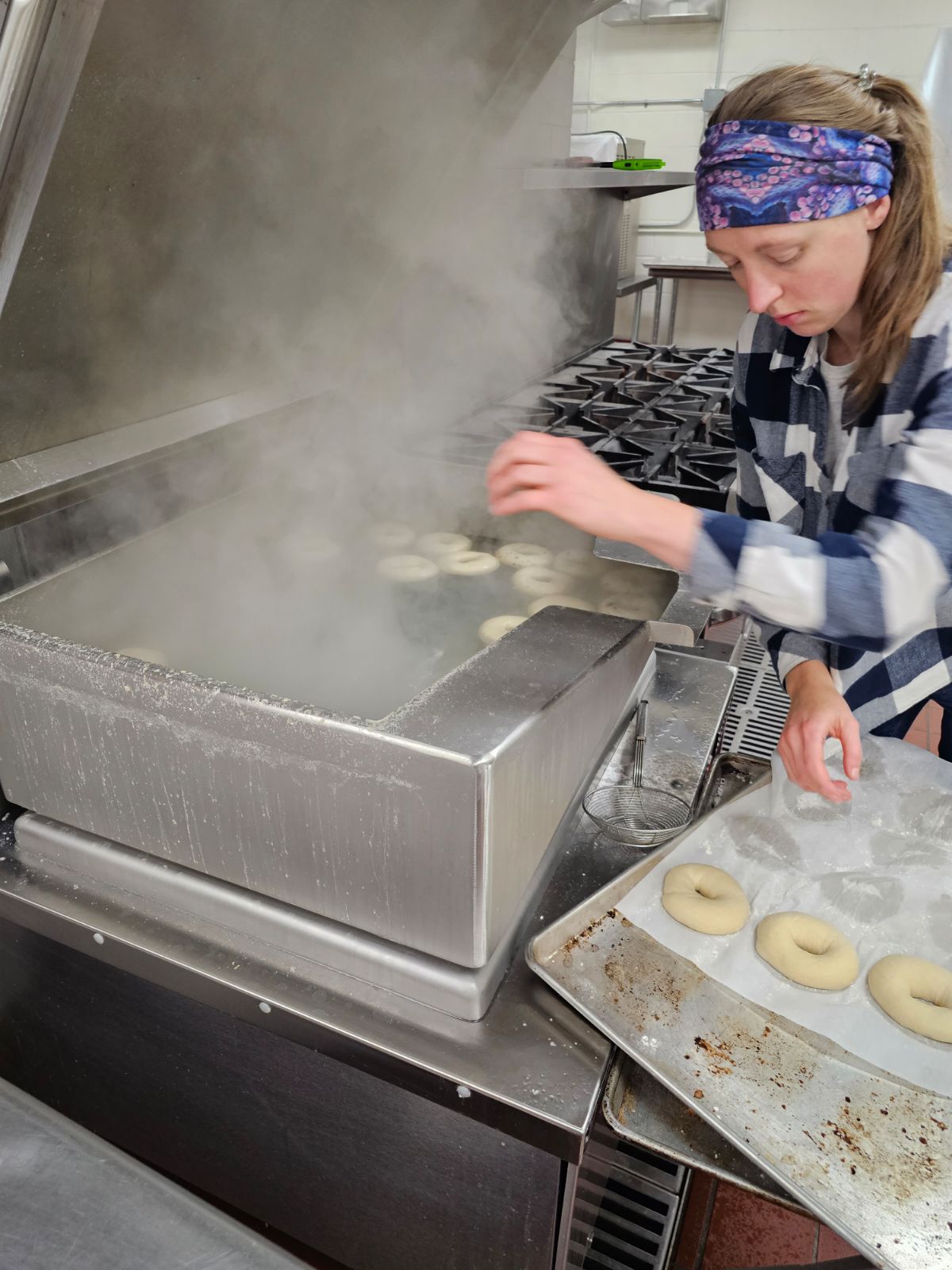 A person wearing a plaid shirt and a colorful headband is boiling bagels in an industrial kitchen. They are placing bagels into a large steaming pot on the stovetop. A tray with unboiled bagels is on the counter beside them.