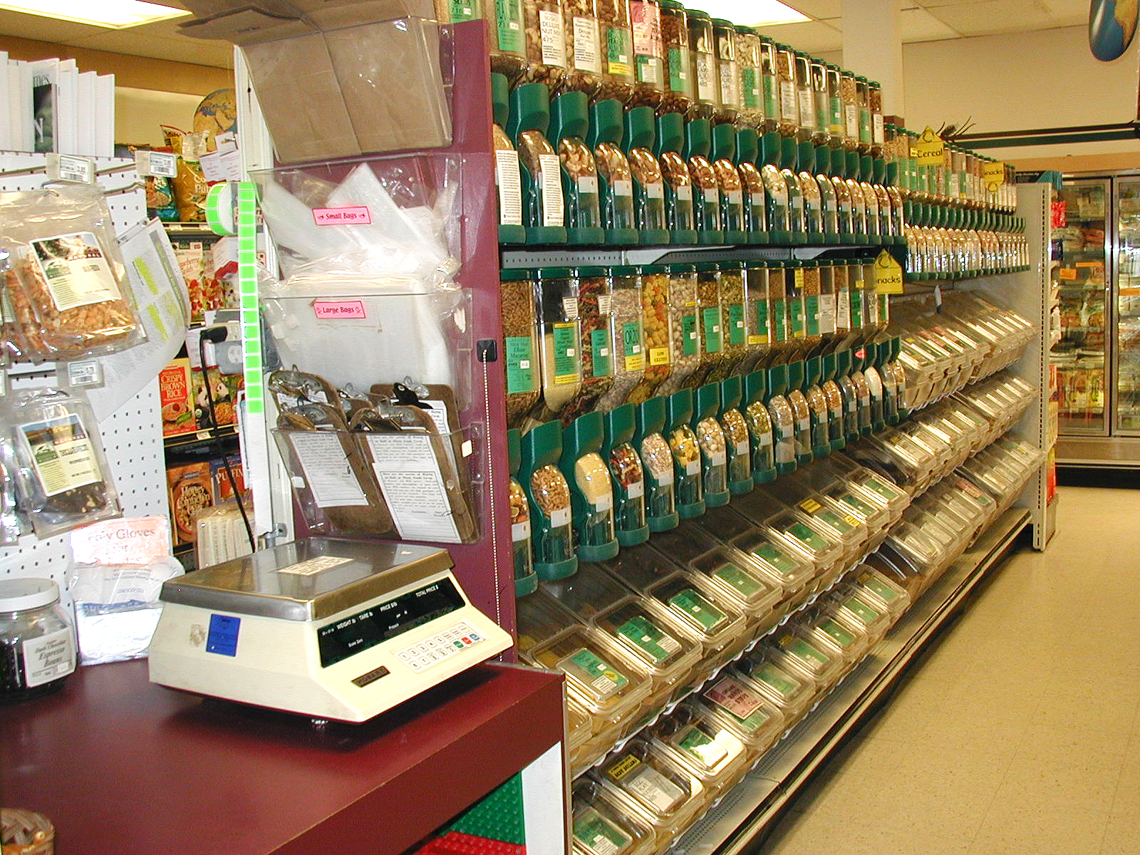 In the Whole Foods Coop Duluth MN, the bulk food section features various containers of grains and legumes neatly displayed on a shelf. The labeled containers dispense products into bags below. A digital scale is conveniently situated on a counter in the foreground, adjacent to the shelf.