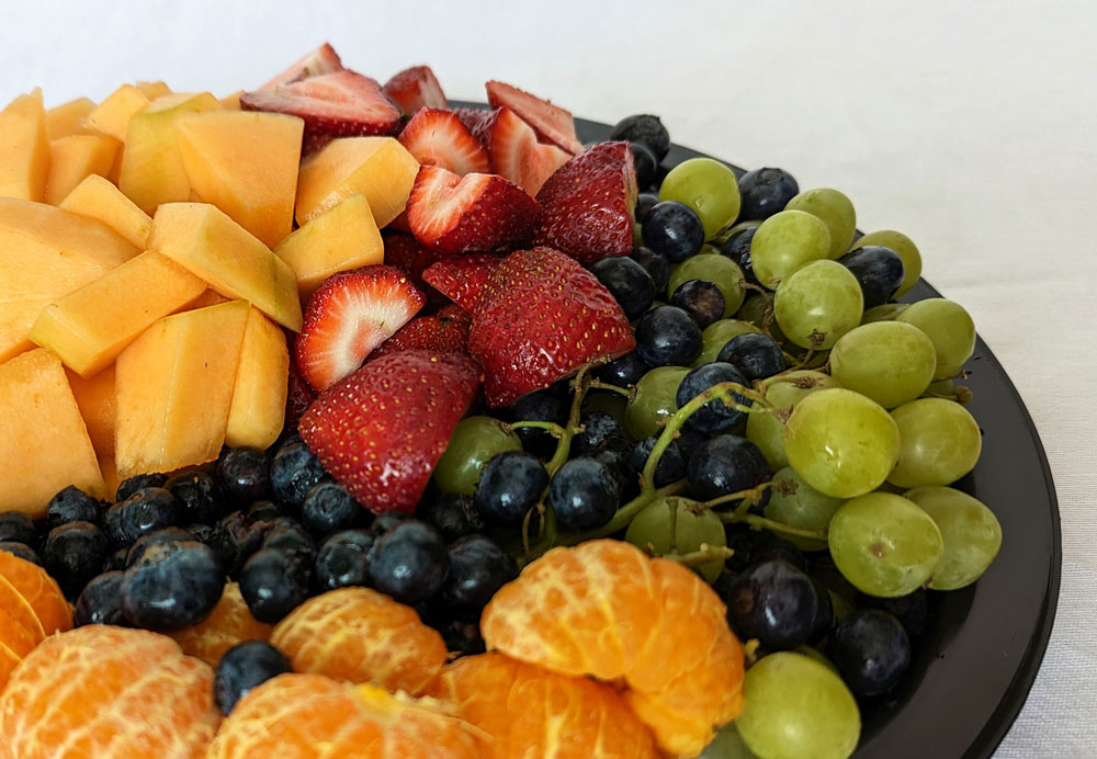 A black plate filled with a variety of colorful fruits, including sliced cantaloupe, strawberries, green grapes, blueberries, and peeled orange segments, is presented on a white tablecloth. The fruits are arranged in separate, vibrant sections—a delightful addition to the Whole Foods Coop Deli Menu.