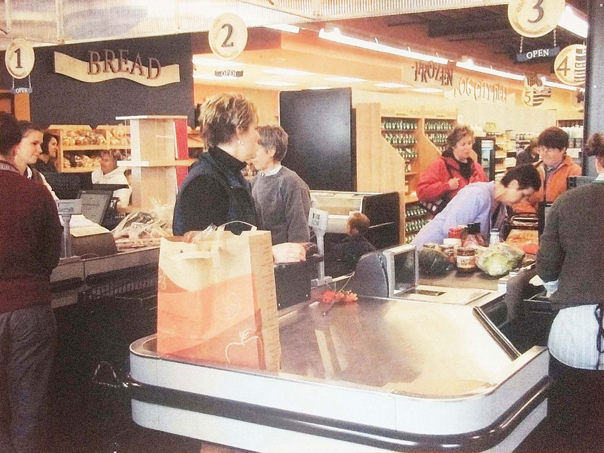 People are waiting in line at the checkout counters in Whole Foods Coop Duluth MN. They are purchasing various items, including produce. Signage for different sections like "Bread" and "Frozen Food Center" is visible. The atmosphere appears busy but orderly.