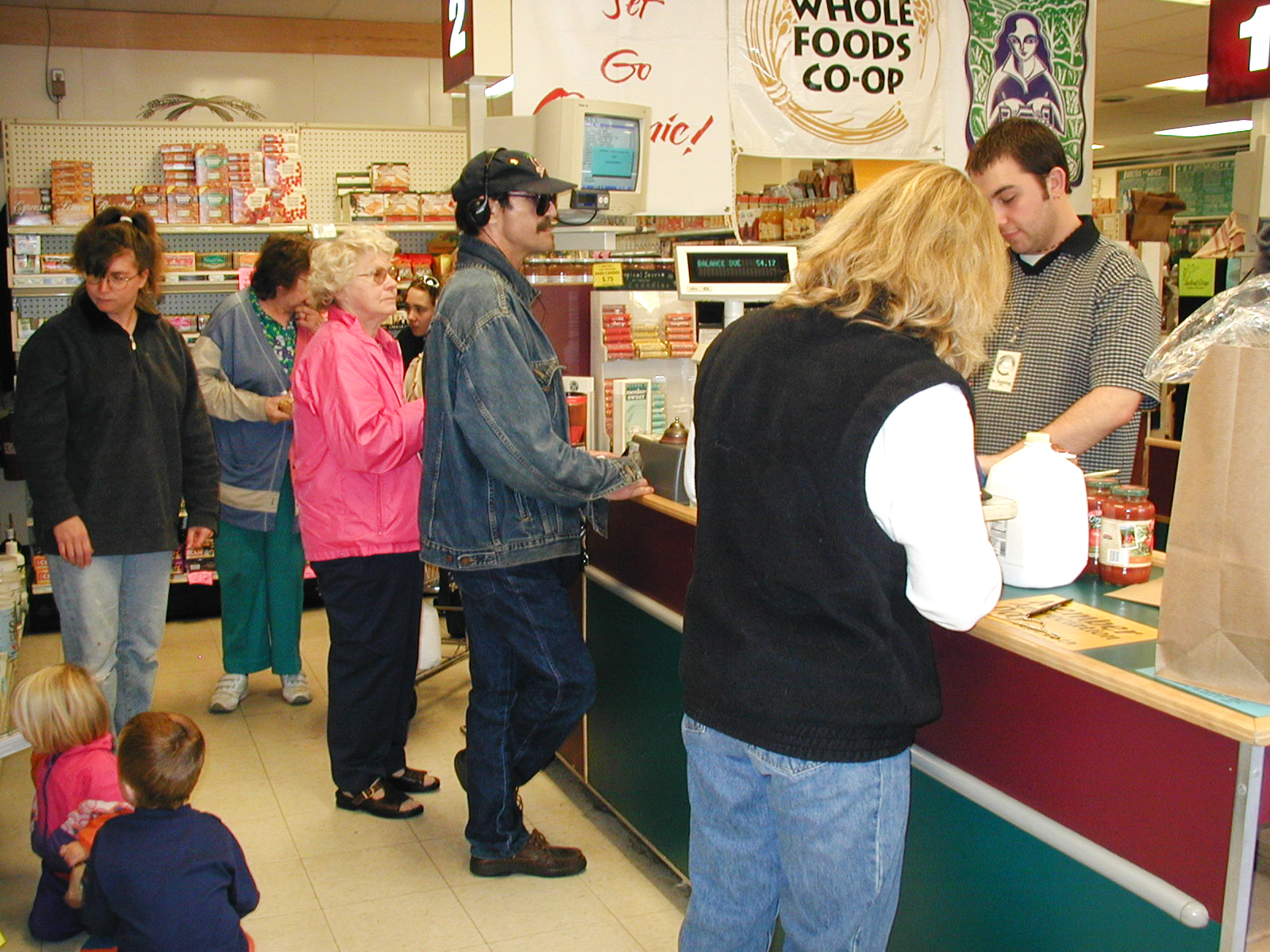 People are standing in line at the checkout counter of Whole Foods Coop Duluth MN. A cashier is scanning items while engaging with a customer in a blue shirt. Shoppers, including children, are waiting or browsing in the background. Various products are visible on the shelves.