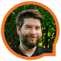 A bearded man with short, dark hair is smiling at the camera. He is wearing a black shirt and is framed by a circular orange border. The background is filled with green, leafy foliage.
