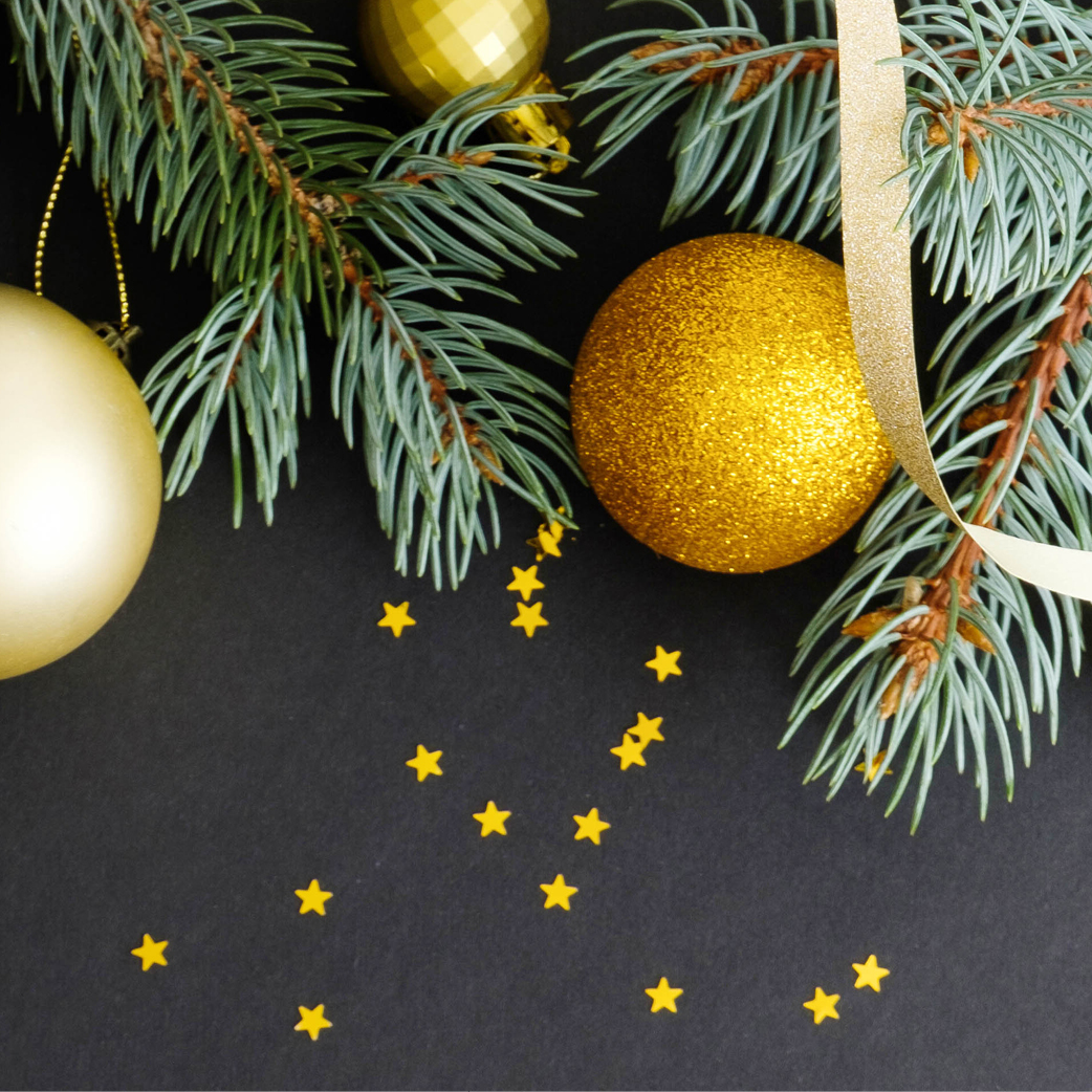 A holiday display featuring gold Christmas ornaments and evergreen branches against a dark background, with sprinkles of small gold star-shaped confetti scattered below. A gold ribbon is also entwined among the branches and ornaments, setting a perfect scene for Christmas Eve.