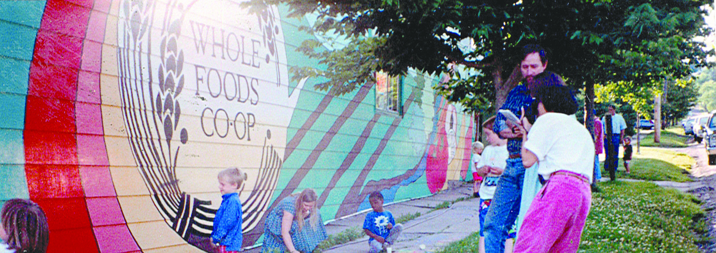 A colorful mural on the side of a building reads "Whole Foods Coop Duluth MN". Adults and children are gathered around the mural, some playing and some conversing. The scene is vibrant with greenery in the background and a sunny atmosphere.