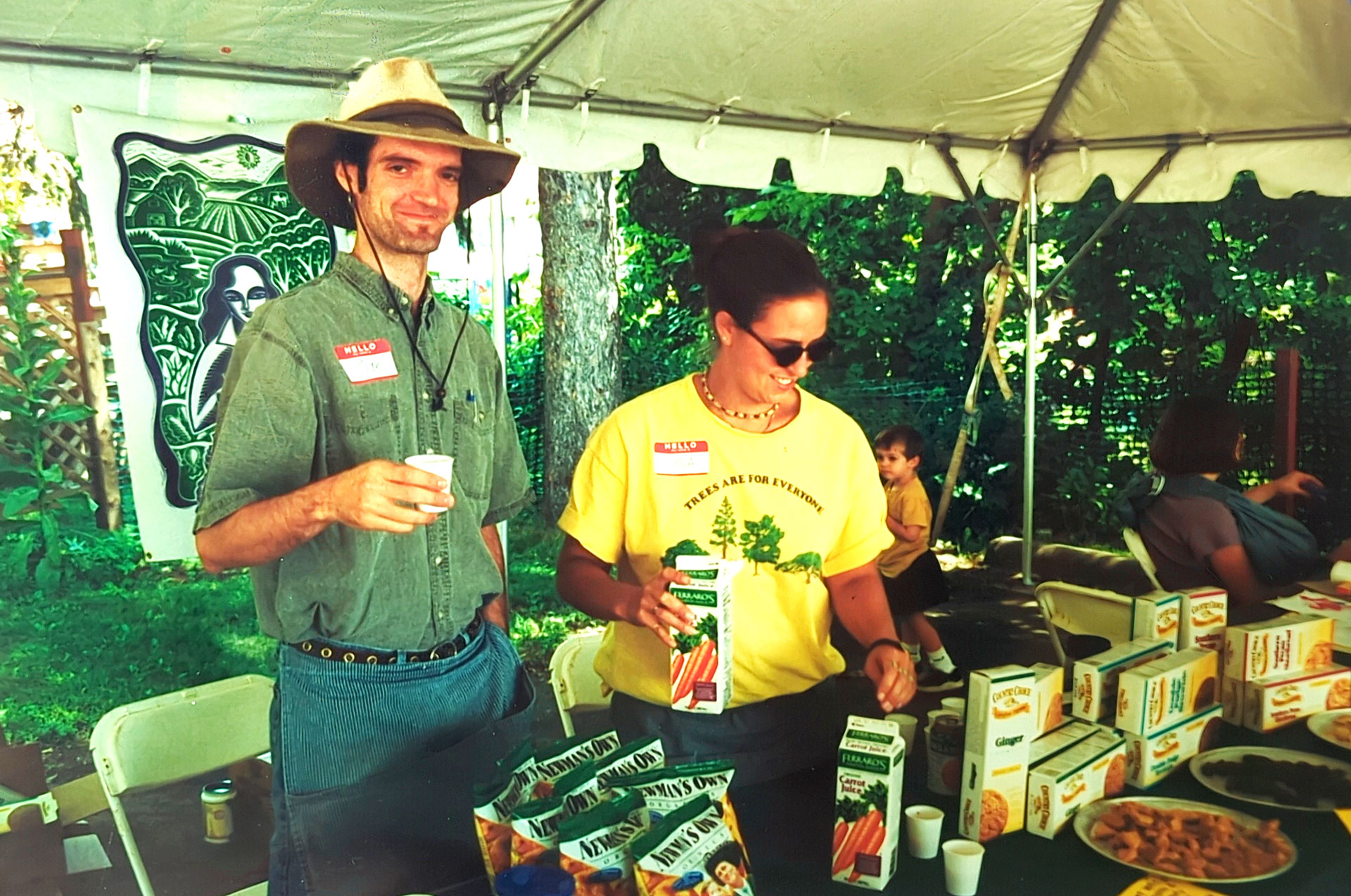 A man and a woman stand under a canopy at an outdoor Whole Foods Coop event in Duluth, MN, each wearing name tags and handling containers of organic cereal. The man has a wide-brimmed hat and a cup in hand, the woman wears a yellow shirt with a graphic, and a child is in the background.