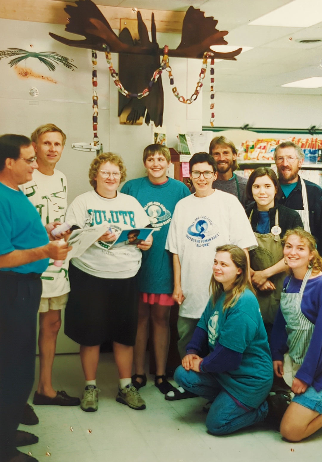A group of nine adults, casually dressed, pose together in a room decorated with a moose head and colorful chains. Some are wearing event T-shirts from the Whole Foods Coop Duluth MN, and one person holds a magazine. They appear cheerful and are positioned in both standing and crouching stances.