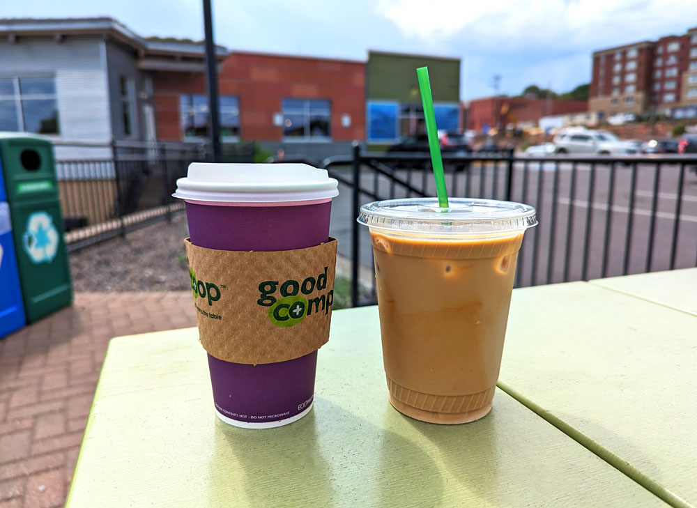 A hot coffee in a purple paper cup with a sleeve and a cold iced coffee in a clear plastic cup sit on a green outdoor table in front of a building. A recycling bin and parked cars are visible in the background on a cloudy day.