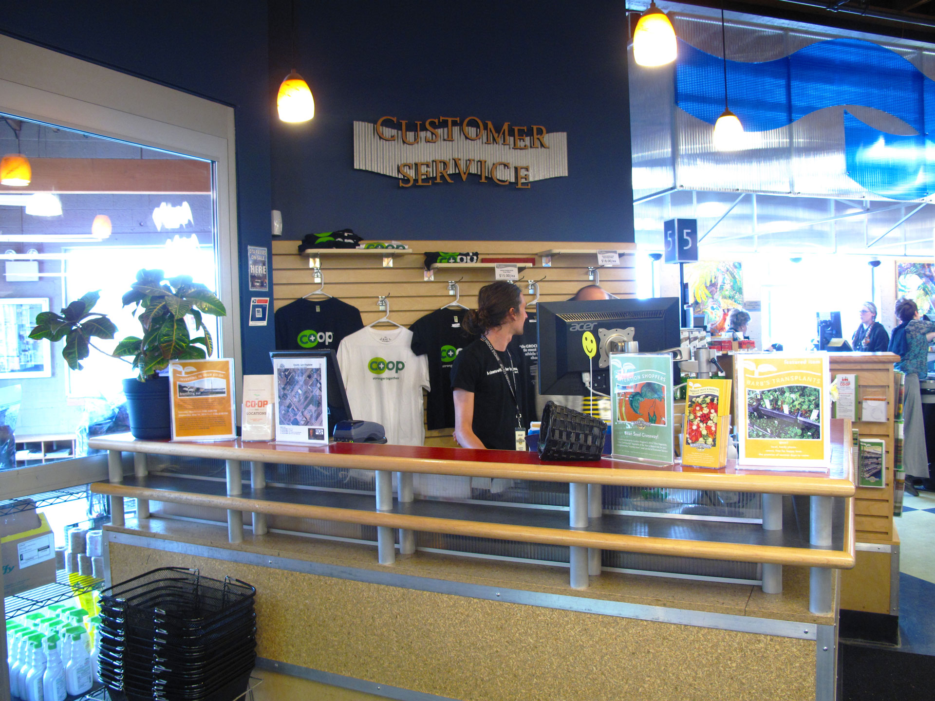A customer service desk at Whole Foods Coop in Duluth, MN features various brochures and flyers displayed. Behind the counter, a uniformed person stands near a computer. Co-op branded t-shirts are hung on the wall, and potted plants decorate the counter.