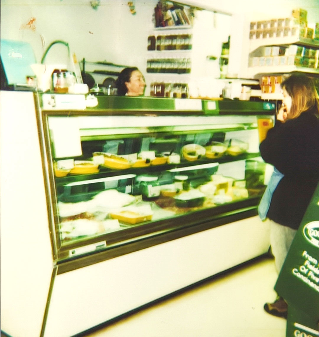 A woman stands at the counter of a grocery store or deli, engaging in conversation with a staff member behind the display case filled with various cheeses and deli items. Shelves stocked with jars and packaged goods are visible in the background, reminiscent of Whole Foods Coop Duluth MN.