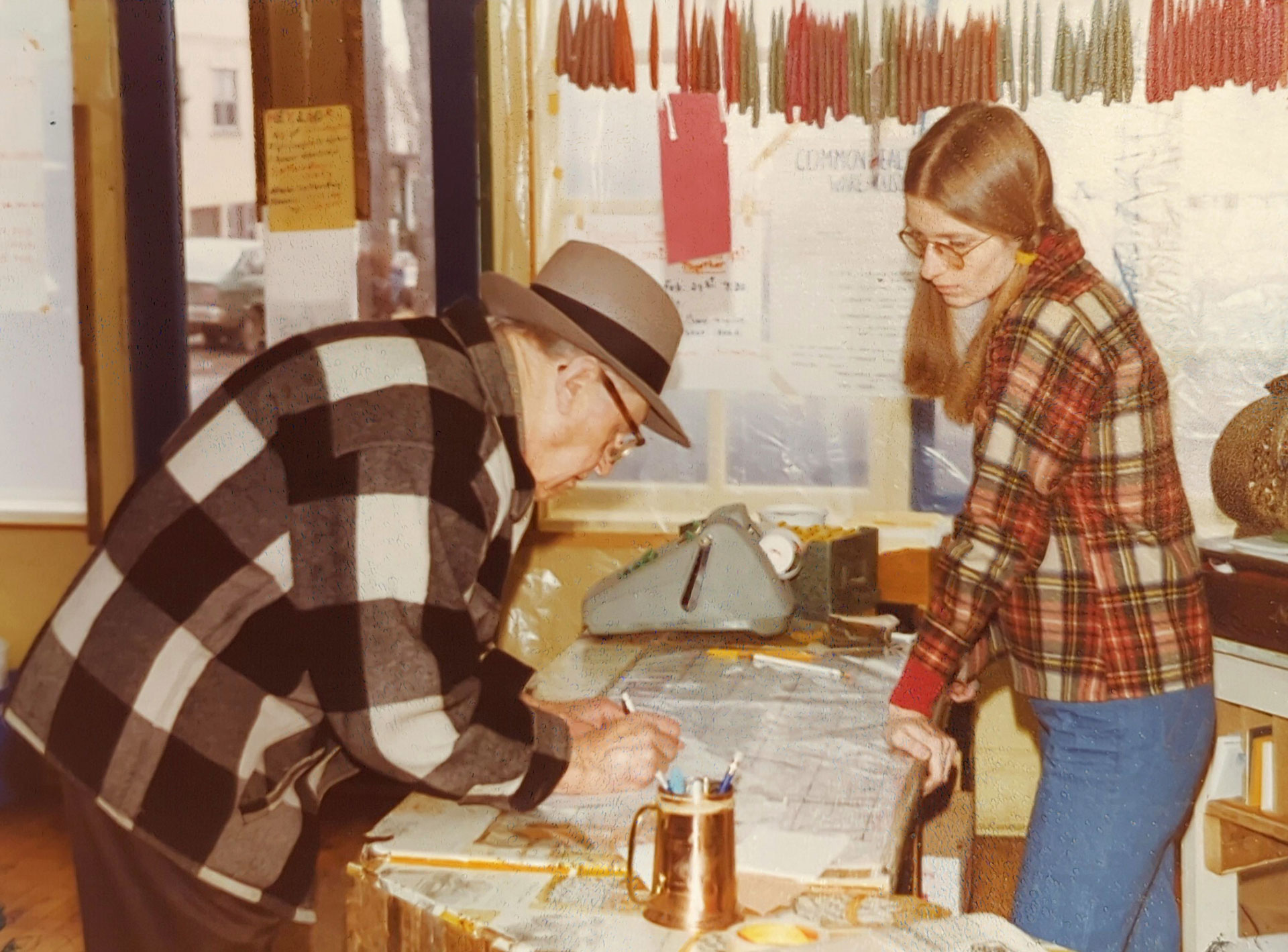 An elderly man in a checkered coat and hat leans over a table studying a document, while a young woman with long hair and glasses, wearing a plaid jacket, stands beside him, also looking at the document. They are in a room with notes pinned on the wall at Whole Foods Coop Duluth MN.