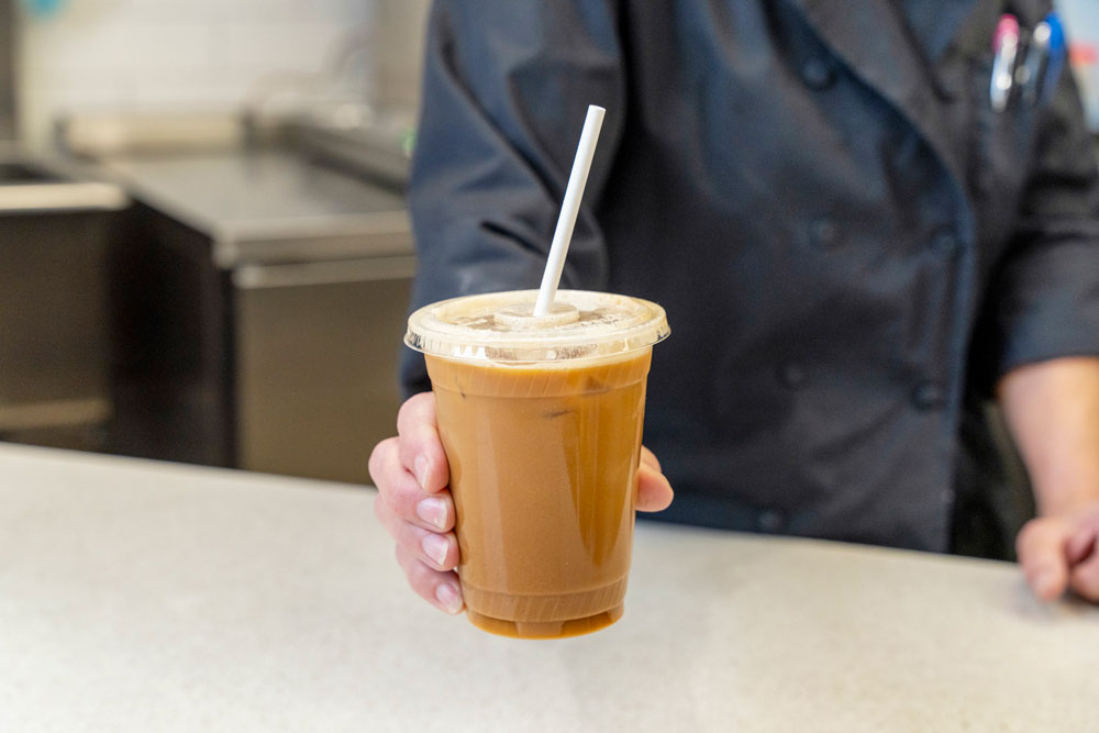 A person in a dark blue chef's uniform holds out a plastic cup filled with iced coffee topped with a lid and a white straw. The background features a kitchen countertop and a partially blurred sink area, reminiscent of the offerings on the Whole Foods Coop Deli Menu.