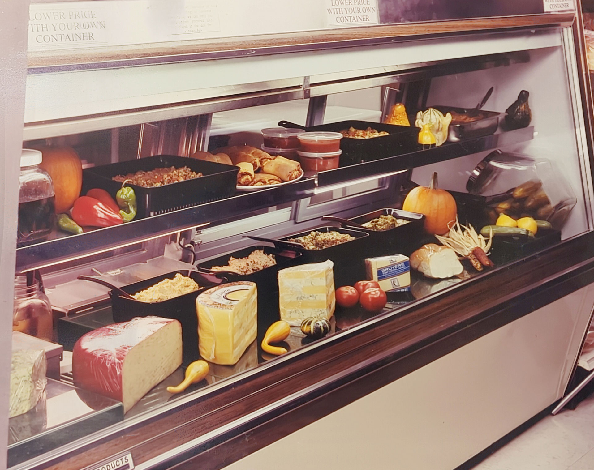A refrigerated deli display at the Whole Foods Coop in Duluth, MN features cheeses, cured meats, vegetables, and various prepared dishes in black trays. Items include pumpkins, bell peppers, tomatoes, and a variety of cheeses. A sign above reads "Lower price when you bring your own container.