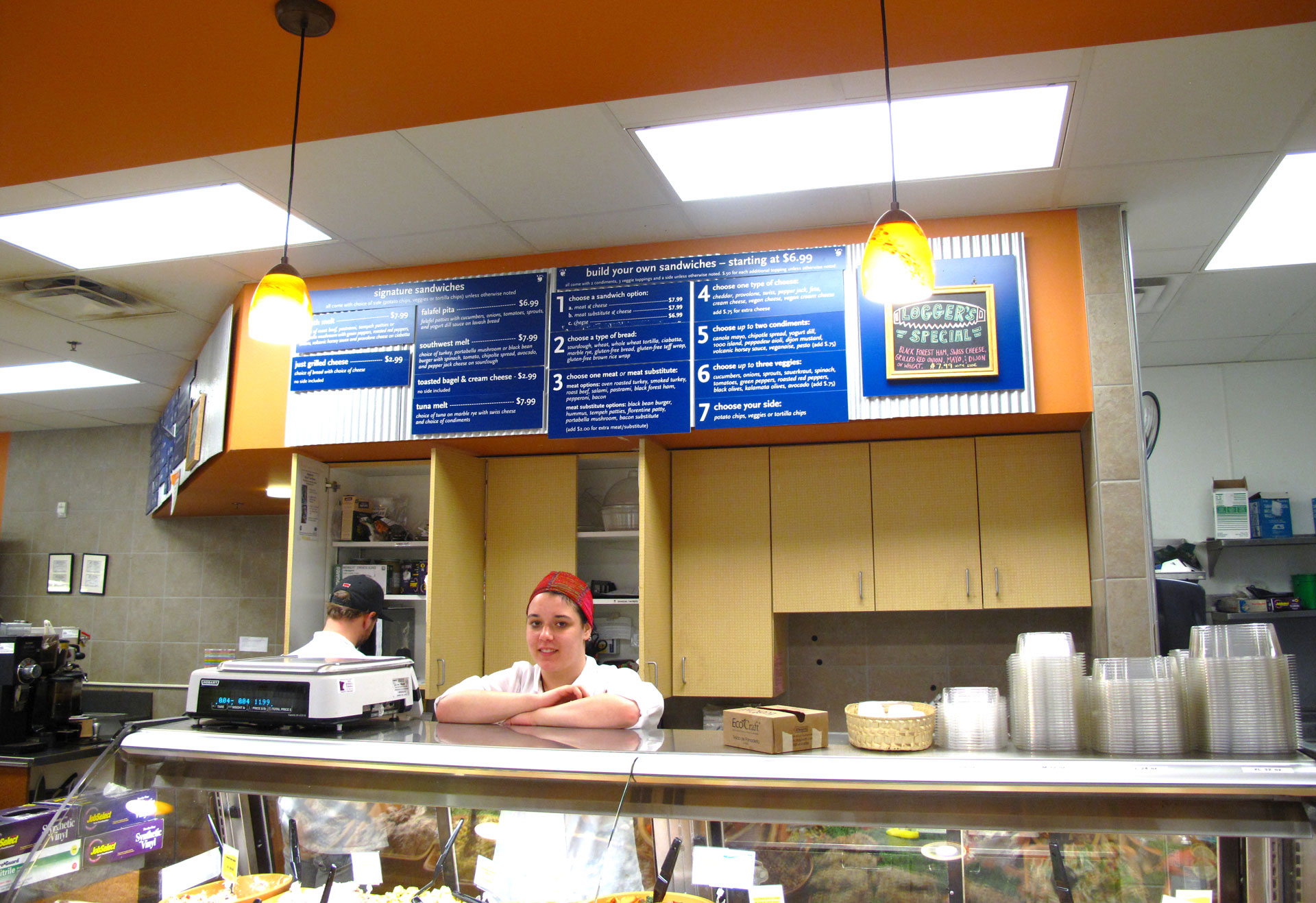 A person wearing a red cap and white uniform stands behind a counter in a sandwich shop at Whole Foods Coop Duluth MN. The shop has a menu displayed on the wall behind them and various food items on the counter. Another person is visible working in the background.