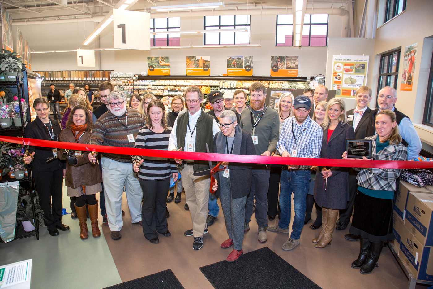 A group of people, including officials and store employees, gather at Whole Foods Coop Duluth MN for a ribbon-cutting ceremony. They are standing behind a large red ribbon, some holding the scissors. The store aisles are visible in the background with products on the shelves.