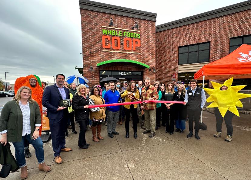 A large group of people stands in front of the Whole Foods Coop Duluth MN store for a ribbon-cutting ceremony. The group includes individuals in business attire, as well as people in costumes, such as a sun and a carrot. A man and woman hold giant scissors to cut the red ribbon.