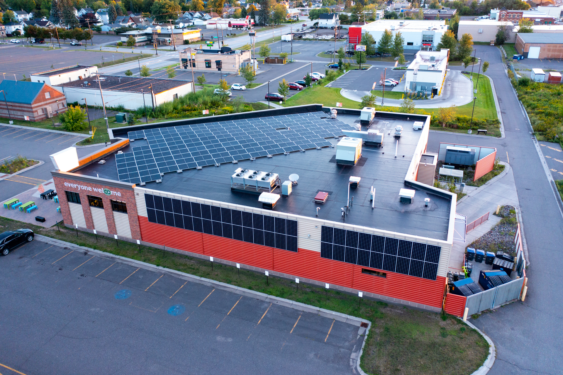 Aerial view of a modern Whole Foods Coop Duluth MN commercial building with solar panels installed on the roof. The building is surrounded by parking lots and other commercial structures. The words "everyone welcome" are displayed on one side of the building. Trees and greenery are in the background.