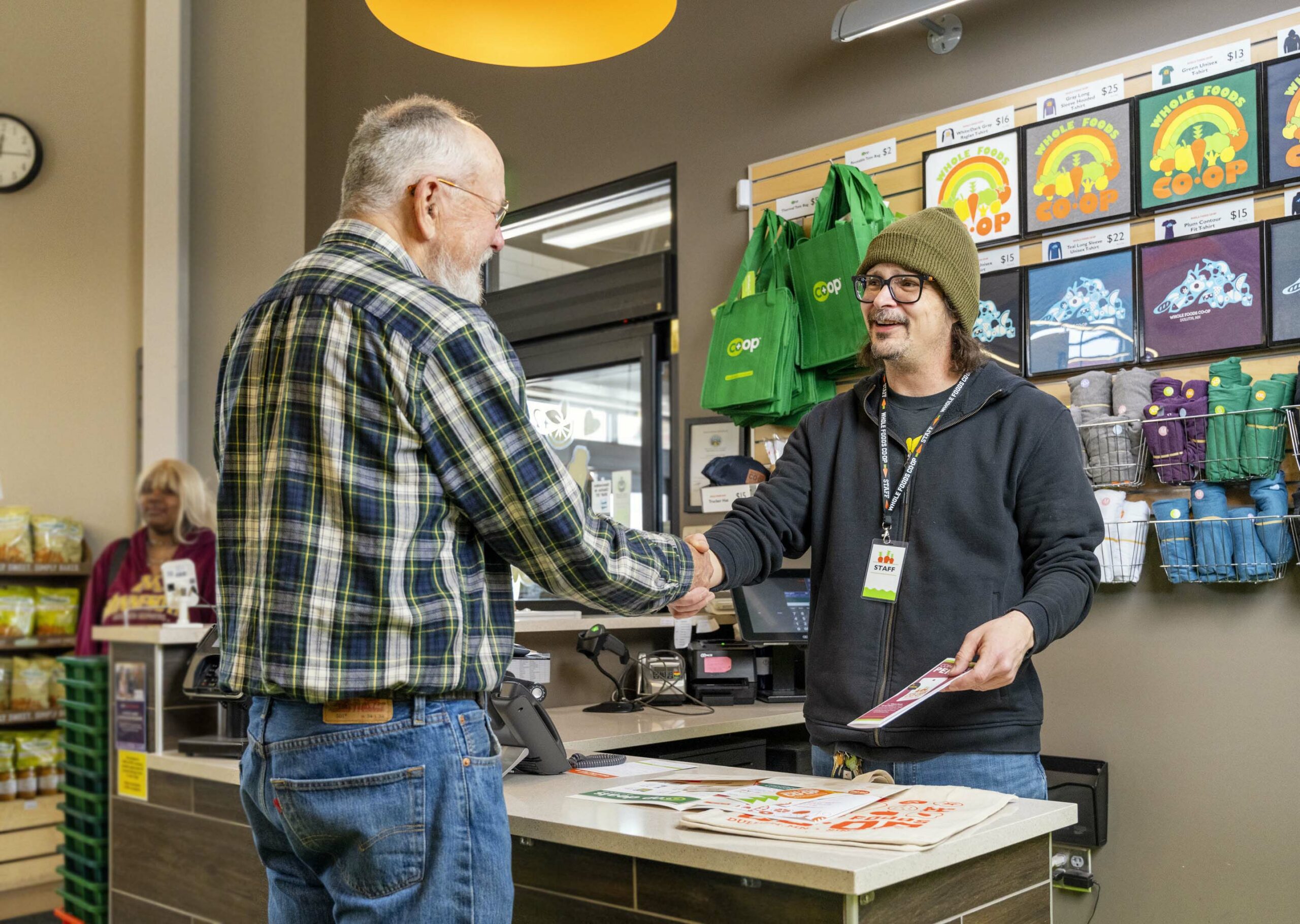 A man in a plaid shirt and a man in a beanie and glasses are shaking hands across a counter at the Whole Foods Coop in Duluth, MN. Shelves behind them display colorful products and reusable bags.