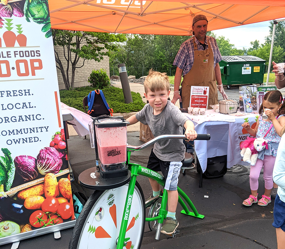 A young boy pedals on a stationary bike powering a blender at a market stall. The stall, covered by an orange tent, is promoting locally grown organic foods from Whole Foods Coop Duluth. An adult in an apron stands behind a display of vegetables and samples, while another child nearby holds a toy unicorn.