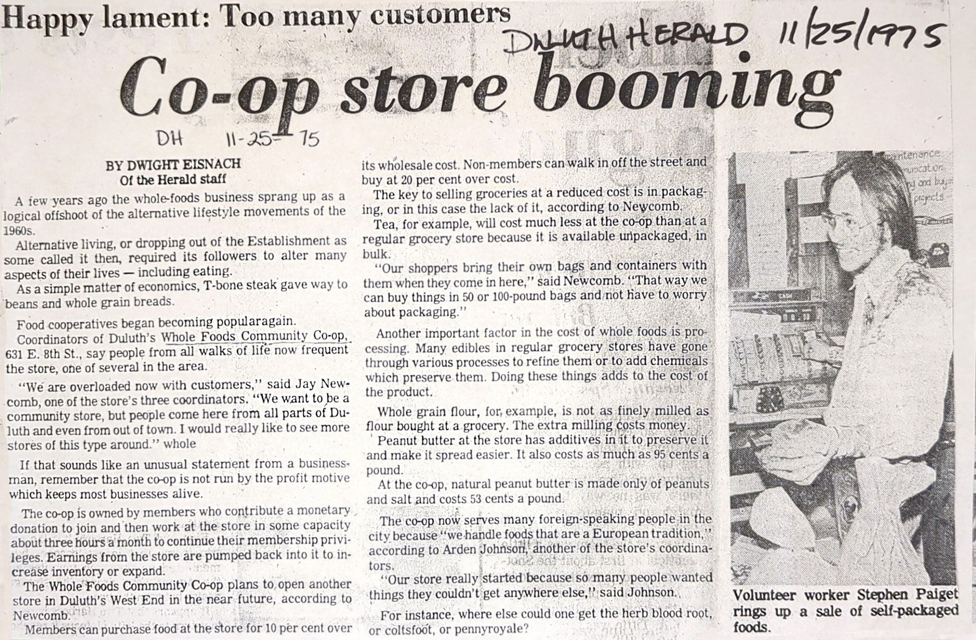 A newspaper clipping titled "Co-op store booming" with handwritten notes indicating the article is from the Duluth Herald, dated 11/25/75. The article discusses the success of Whole Foods Coop Duluth MN. On the right, a volunteer named Stephen Paiget is seen stocking shelves.