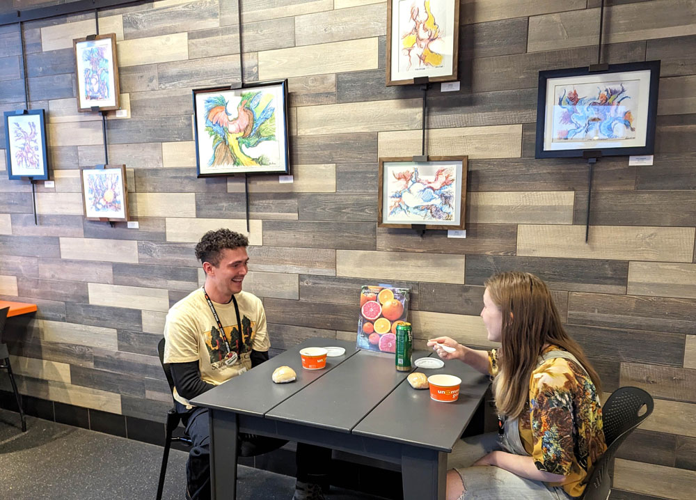 Two people sit at a small table in a cozy café, enjoying bowls of soup from the Whole Foods Coop in Duluth, MN. They are smiling and engaged in conversation. The café's wood-paneled wall is adorned with colorful, abstract artwork in black frames, adding to the warm and inviting atmosphere.