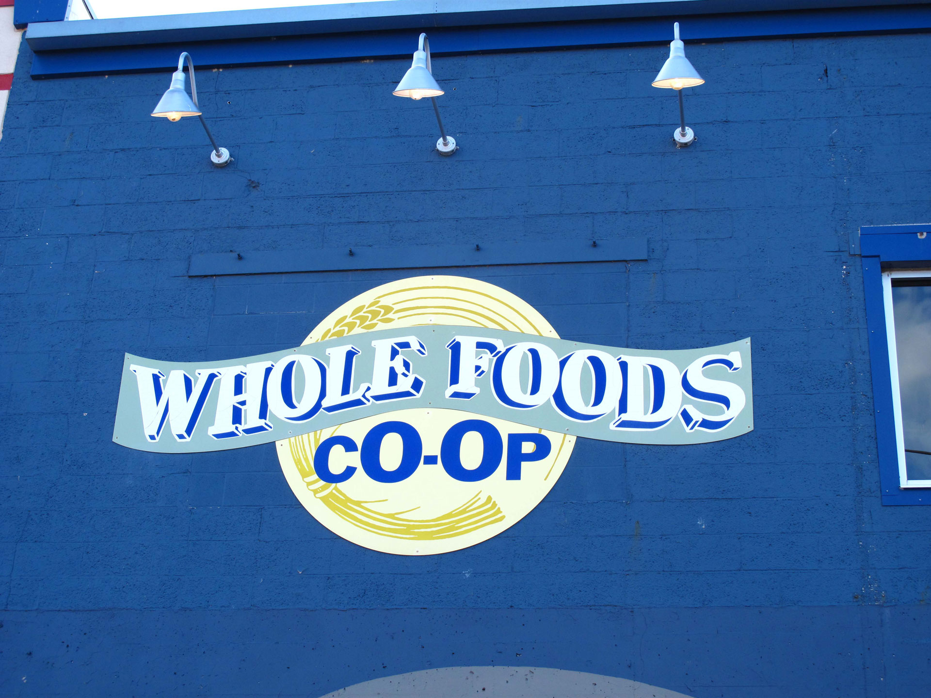 A blue storefront wall with three industrial light fixtures illuminates a circular sign that proudly reads "Whole Foods Coop Duluth MN" in blue and yellow letters, accompanied by a wheat graphic. A small portion of a window frame is visible on the right side.