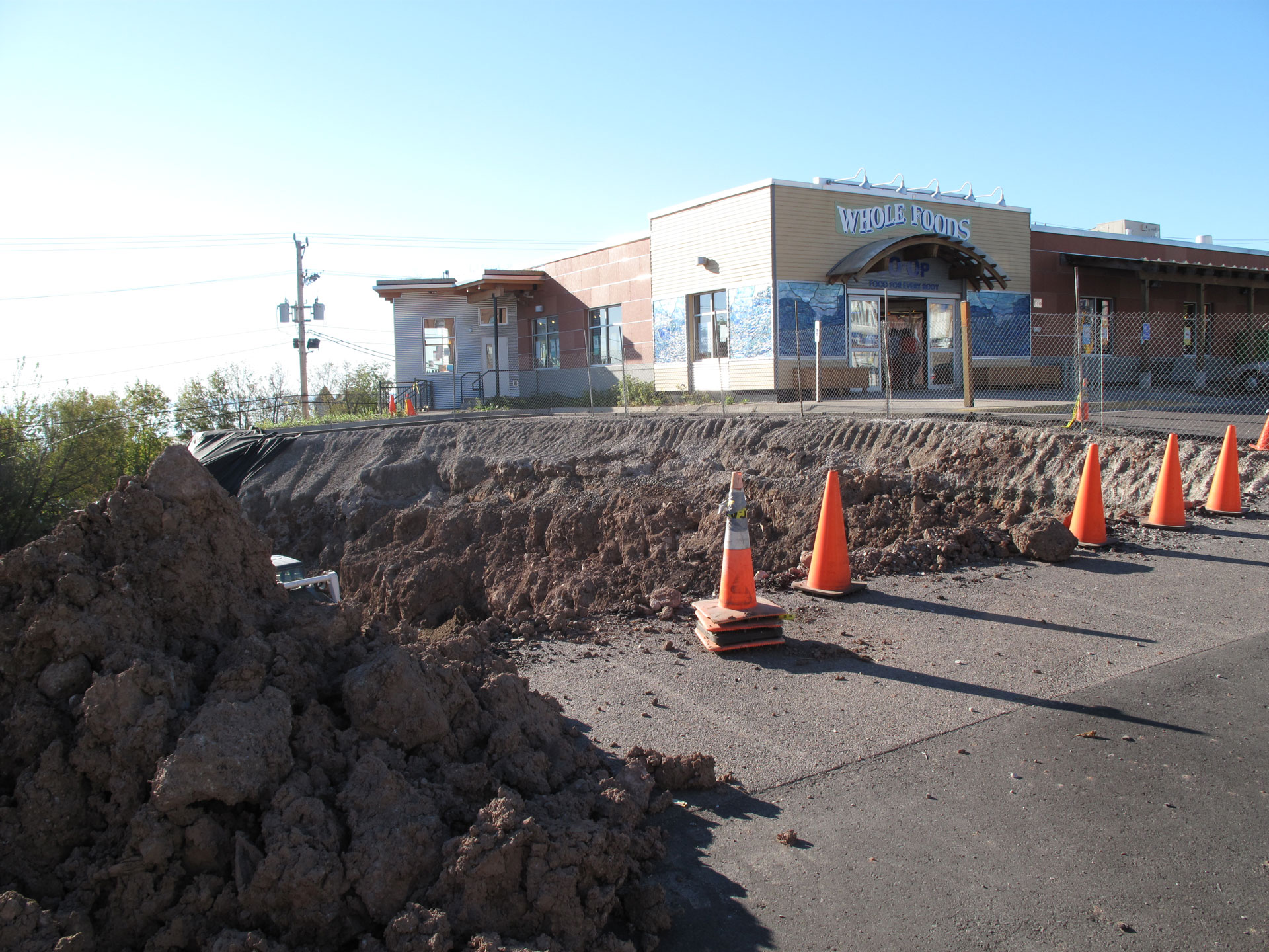 Construction site in front of the Whole Foods Coop in Duluth, MN. The area is fenced off with orange cones and dug up soil piled beside a trench. The store's entrance is visible behind the barriers, and the sky is clear.