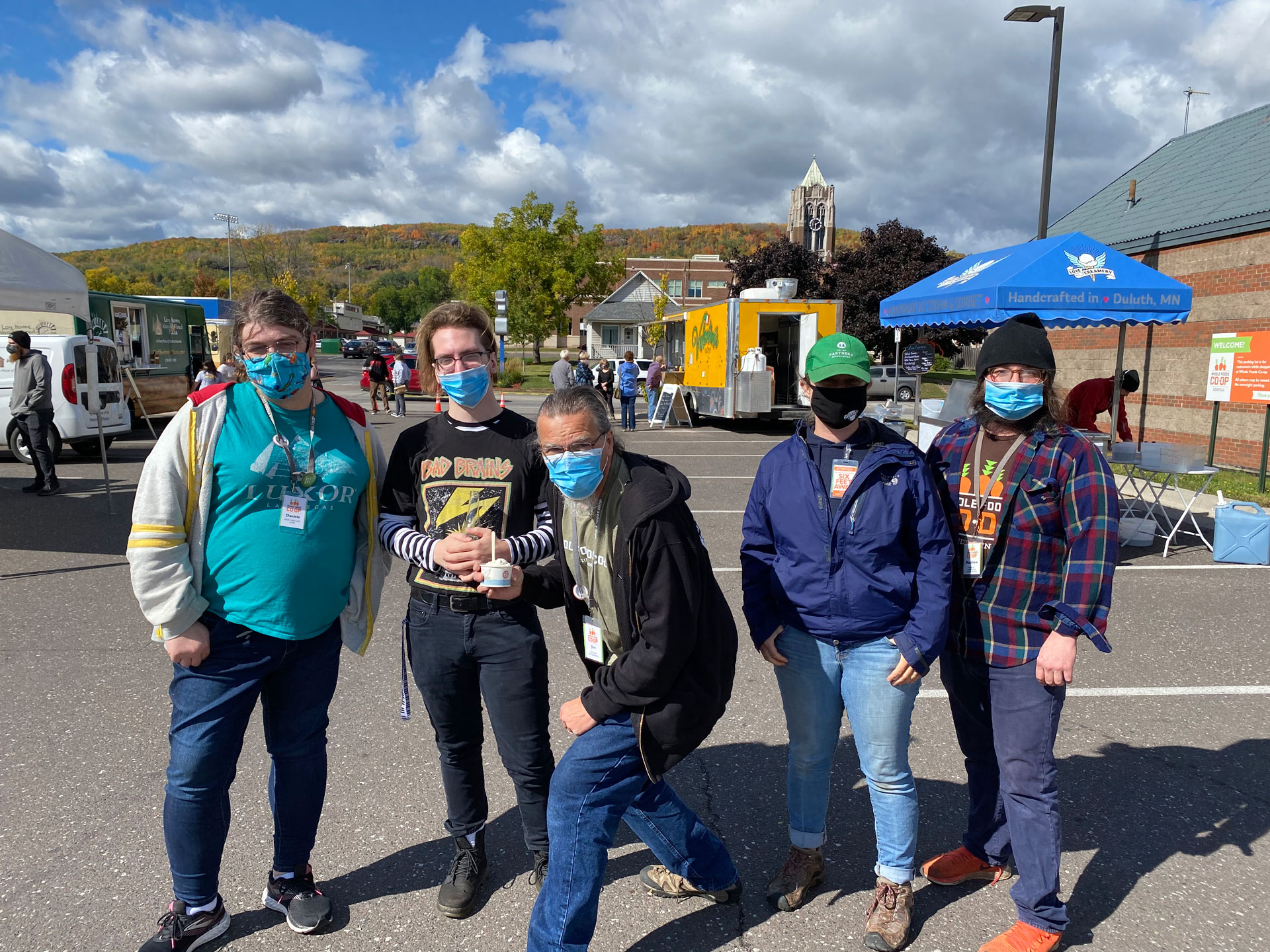 Five people, all wearing face masks, stand outdoors in a parking lot with food trucks and buildings in the background. They are grouped together, with some posing with arms around one another. The sky is partly cloudy, with trees and a hill visible in the distance near Whole Foods Coop Duluth MN.