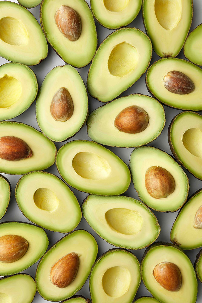 A neatly arranged pattern of halved avocados. Some halves have their seeds intact, while others have empty pits. The vibrant green flesh contrasts with the brown seeds and the dark green, textured skin.